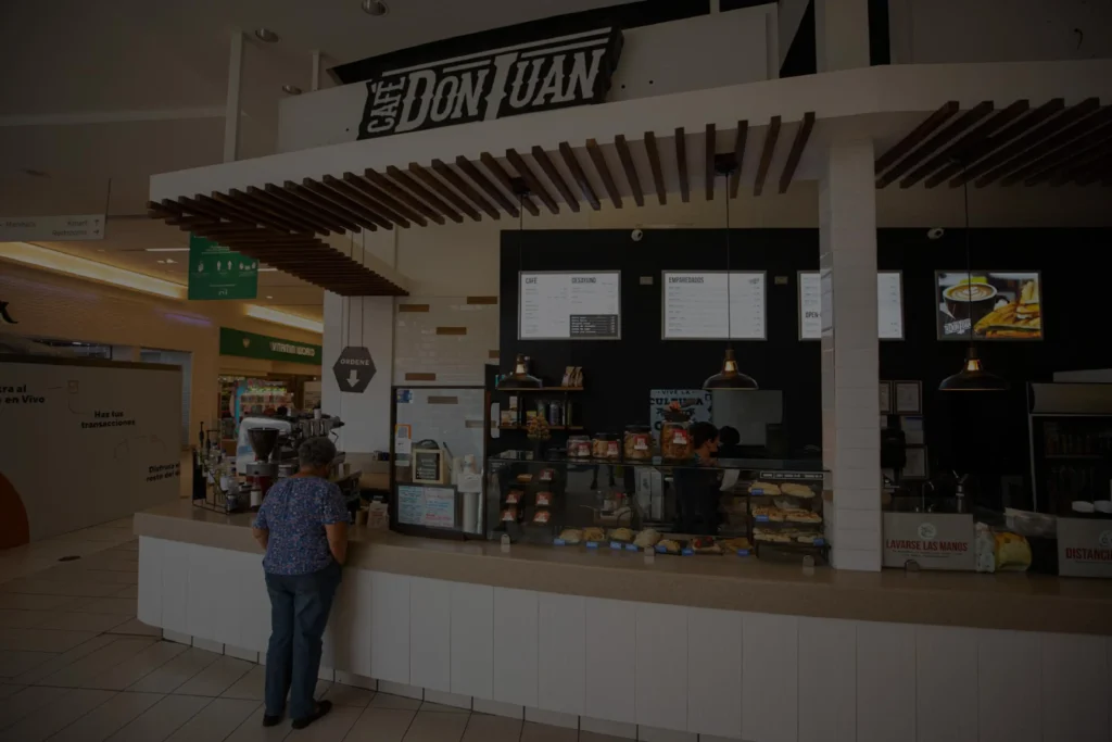 Tienda de Café Don Juan dentro del centro comercial Plaza Rio Hondo con clientes de pie frente al mostrador y vitrinas de productos, mostrada con un filtro oscuro semitransparente.