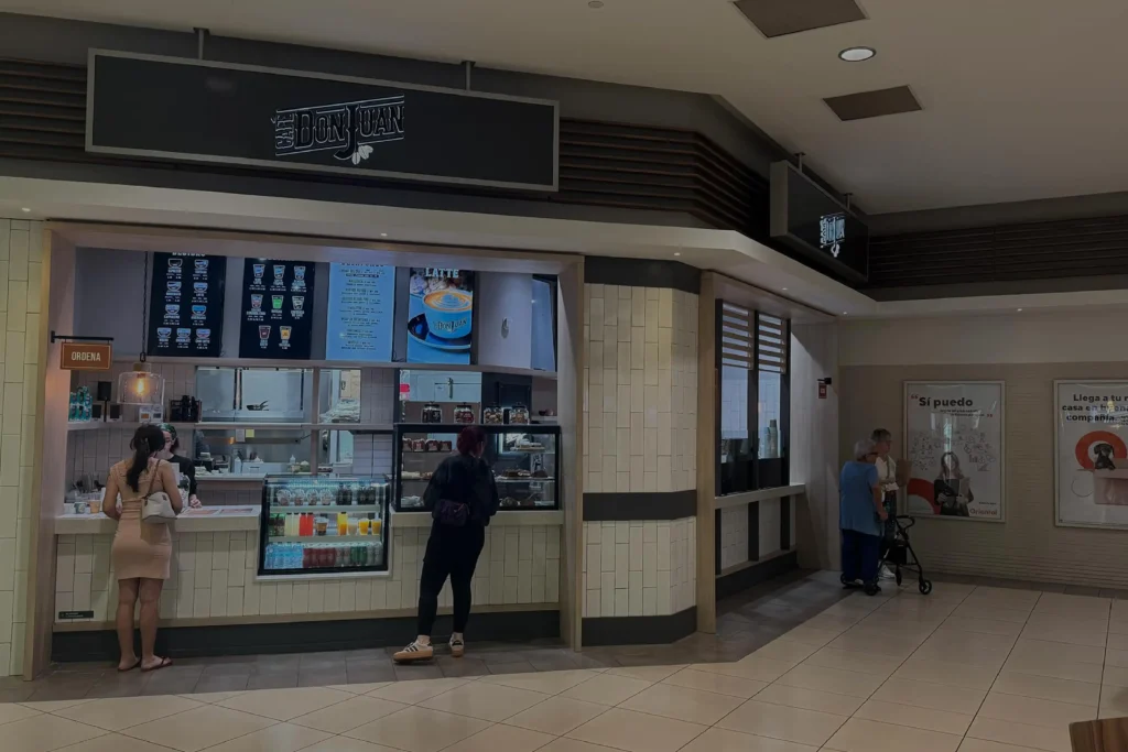 Tienda de Café Don Juan dentro del centro comercial Plaza Carolina con clientes de pie frente al mostrador y vitrinas de productos, mostrada con un filtro oscuro semitransparente.