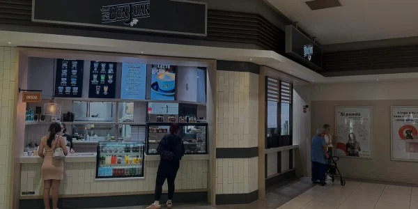 Tienda de Café Don Juan dentro del centro comercial Plaza Carolina con clientes de pie frente al mostrador y vitrinas de productos, mostrada con un filtro oscuro semitransparente.