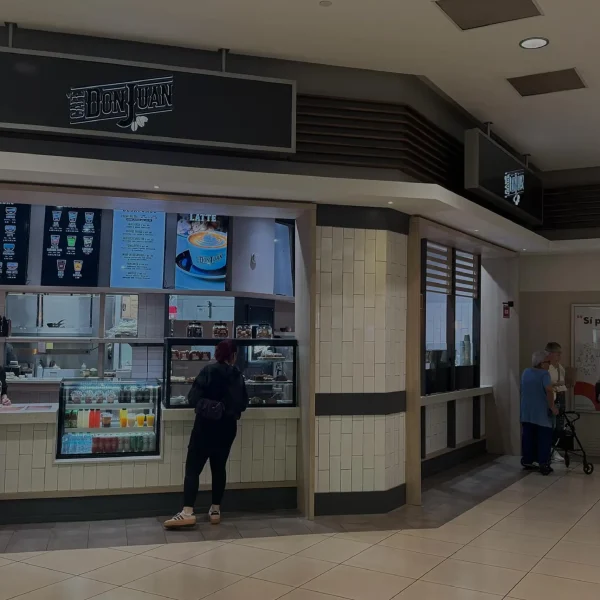 Tienda de Café Don Juan dentro del centro comercial Plaza Carolina con clientes de pie frente al mostrador y vitrinas de productos, mostrada con un filtro oscuro semitransparente.
