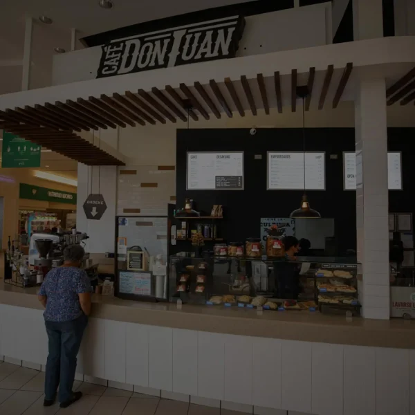 Tienda de Café Don Juan dentro del centro comercial Plaza Rio Hondo con clientes de pie frente al mostrador y vitrinas de productos, mostrada con un filtro oscuro semitransparente.